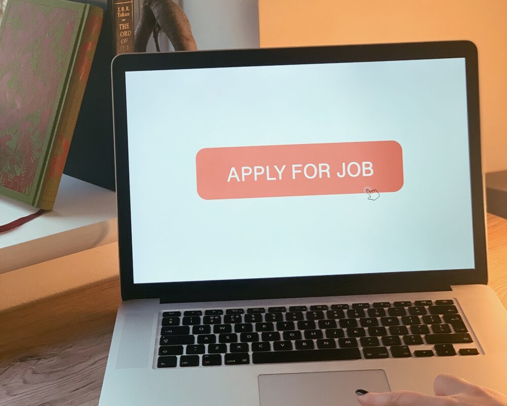 Image shows a laptop on a wooden desk with a hand clicking an orange 'Apply for Job' button