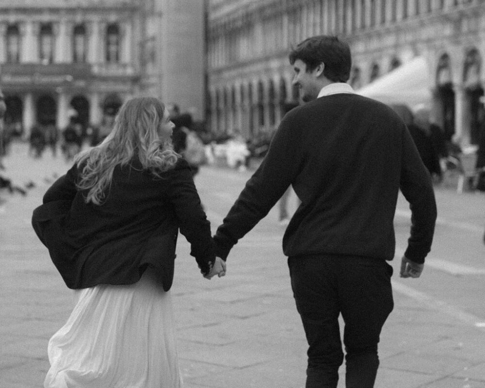Black and white photo of Sophie & Ryan holding hands and running into St Marks Square, Venice