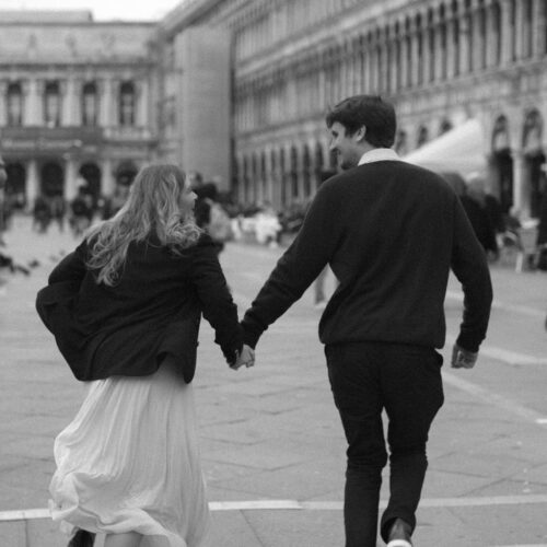 Black and white photo of Sophie & Ryan holding hands and running into St Marks Square, Venice