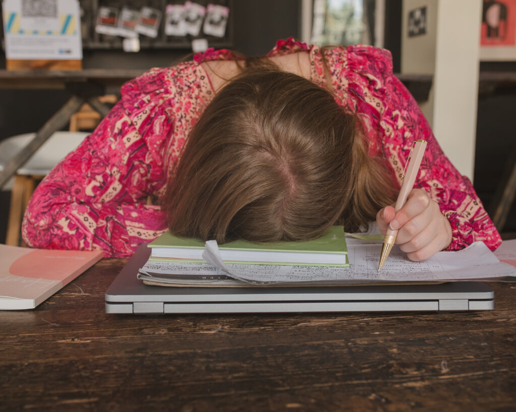 Sophie's head is on a dark wooden table and she is feeling overwhelmed, there are journals, laptop and papers under her head. She is holding a pink pen in her right hand and wearing a fuschia pink 'Happy Feelings' Free People dress 